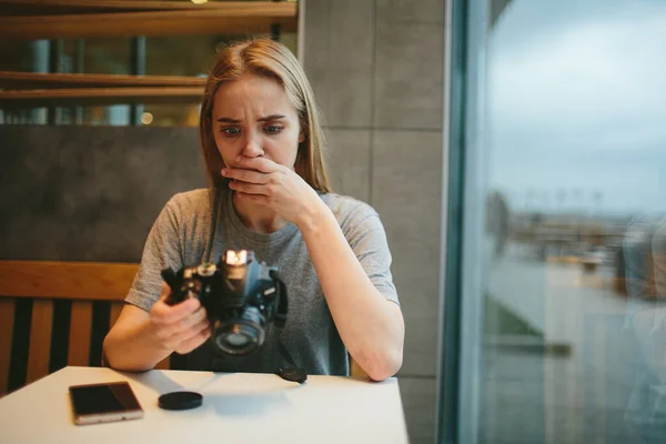 Junge Blondine in einem Café mit einer Kamera. Blickt auf den Bildschirm. Sie ist überrascht und aufgeregt. — Stockfoto