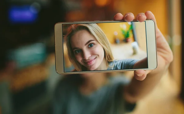 Młoda blondynka w kawiarni. Pokazuje smartfona w aparacie. Na ekranie jej zdjęcie — Zdjęcie stockowe