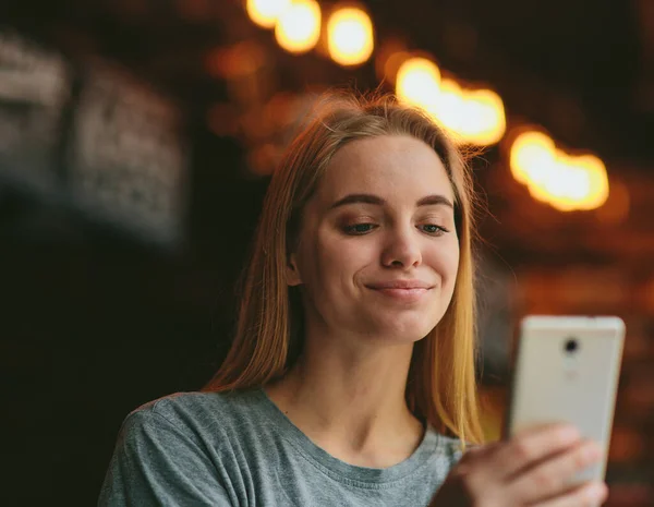 Schöne junge Frau im Café oder Restaurant hält Smartphone in der Hand und schaut es lächelnd an. Fröhliche, glückliche Frau mit modernen Technologien. — Stockfoto