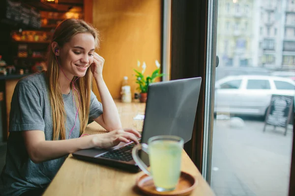在咖啡馆里，年轻的金发姑娘与笔记本电脑一起工作，听音乐。桌上放着一杯绿茶. — 图库照片