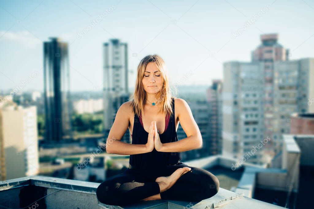 Young woman practices yoga outside. Calm peaceful girl sitting on parapet in lotus position. Her hands at chest level touch each other. City and high-rise buildings on background.