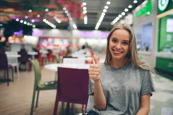 Fröhlich schöne junge Frau zeigt Daumen nach oben in einem Food Court mit unkonzentriertem Hintergrund — Stockfoto