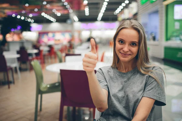 Fröhlich schöne junge Frau zeigt Daumen nach oben in einem Food Court mit unkonzentriertem Hintergrund — Stockfoto