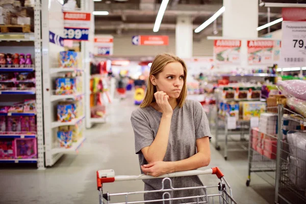 Любопытная женщина в супермаркете. Молодая девушка в магазине с магазинами думает, что купить — стоковое фото