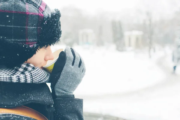 Unga kvinnor klädda i varm hatt rock halsduk och handskar utanför med pappersglas i händerna. Snö på hennes kläder. Hon vill ha en varm drink. Begreppet vintertid. Snöig vinter på bakgrunden. — Stockfoto