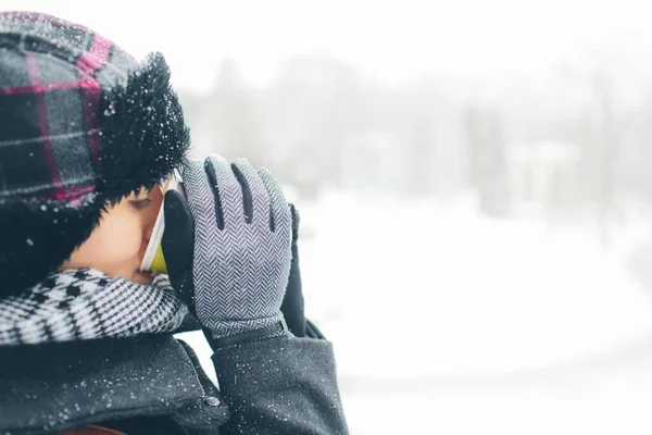 Jeunes femmes vêtues d'un manteau chaud écharpe et gants à l'extérieur avec du verre de papier dans les mains. Neige sur ses vêtements. Elle boit sa boisson. Concept d'heure d'hiver. Hiver enneigé sur fond . — Photo