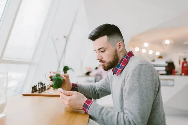 Young bearded man in cafe. He dressed in gray pullover and plaid shirt. Man holds tea and looks at smartphone . Chessboard flower and blurred interior of cafe on background. — Stock Photo, Image