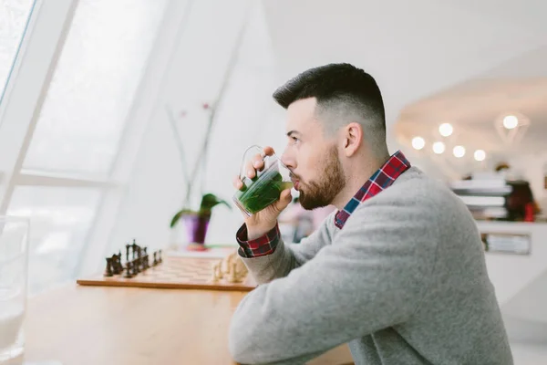 Young bearded man in cafe. He dressed in gray pullover and plaid shirt. Sits at long table near window. Man drinks beverage with green leaves. On table chessboard. Laconic interior. — Stock Photo, Image