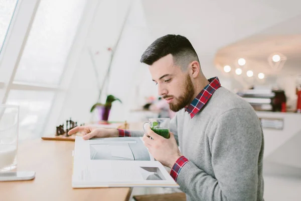 Young bearded man in cafe. He sits at long table near window. Guy hold glass with green leaves on one hand. Another on magazine that man looking at. Blurred background. — Stock Photo, Image