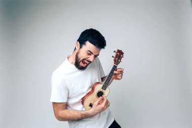 Genç adam arka planda izole edilmiş. Erkek sanatçı ukulele çalıp şarkı söylüyor. Yüksek sesle bağırıyordu. Küçük gitarla çalma pratiği. Stüdyoda tek başına eğlen.