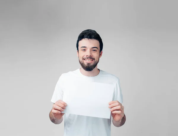 Young man isolated over background. Bearded guy in white shirt hold blank page in hands and smile on camera. Alone in studio. — Stock Photo, Image