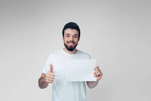 Young man isolated over background. Positive cheerful guy hold white blank page and hold big thumb up. Young hipster in shirt alone in studio. — Stock Photo, Image