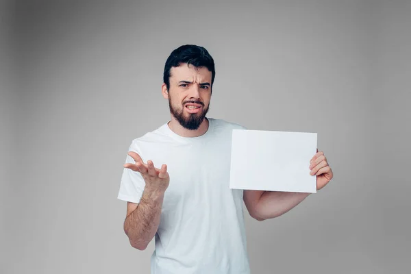 Young man isolated over background. Guy hesitate on camera. Emotional expression in studio. Hold white blank page in hands. Brunette feel disgution. — Stock Photo, Image