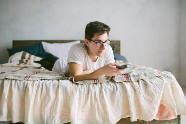 Evdeki yatakta otururken uzaktan kumanda kullanan adam. Televizyon izleyen mutlu, çekici bir genç adam. Yüzünde insani duygular