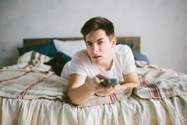 Хлопець використовує пульт дистанційного керування, сидячи на ліжку вдома. Щасливий привабливий молодий чоловік дивиться телевізор. Людські емоції на її обличчі . — стокове фото