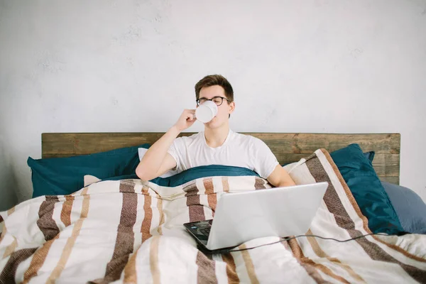 Przypadkowy młody człowiek za pomocą laptopa w łóżku w domu i picia kawy — Zdjęcie stockowe