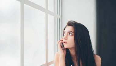 Pencerede beyaz tişörtlü, güneş ışığına boğulmuş genç bir kadın akıllı telefon kullanıyor, iletişim kuruyor ve internet ya da sosyal ağlarda bilgi arıyor.