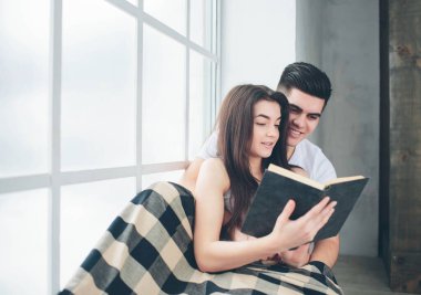 Genç bir adam ve bir kadın pencerenin kenarında oturuyorlar ve güneşli pencerenin yanında kitap okuyorlar..