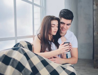 Genç bir adam ve bir kadın sabahleyin yatakta kahve içiyorlar. Evde romantik bir sabah.