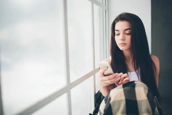 Una mujer joven con el pelo negro en una camiseta blanca en una ventana inundada de luz solar utiliza un teléfono inteligente, se comunica y busca información en Internet o redes sociales — Foto de Stock