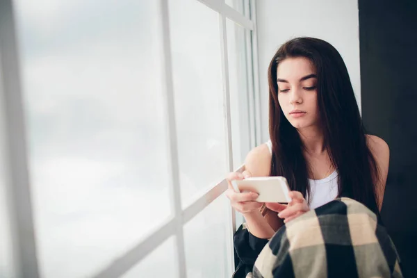 一位穿着白色T恤衫、留着黑头发的年轻女子，站在阳光普照的窗前，用智能手机与人交流，并在网上或社交网络上搜索相关信息 — 图库照片