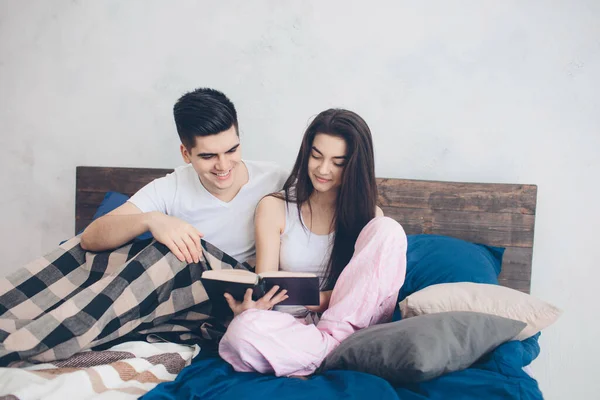 Un hombre y una mujer jóvenes están sentados en la cama y leyendo un libro. Mañana romántica en casa — Foto de Stock