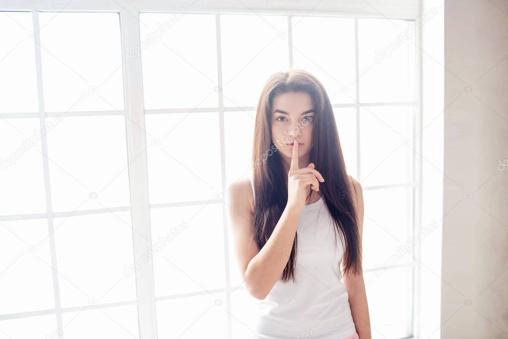 A young woman with black hair in a white T-shirt at a window bathed in sunlight. Morning