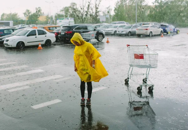 Menina em capa de chuva amarela ficar sozinho sob chuva no estacionamento. Olhe para baixo no asfalto molhado. Muitos carros para trás. Carrinho nojento vazio ao lado. Tempo chuvoso . — Fotografia de Stock