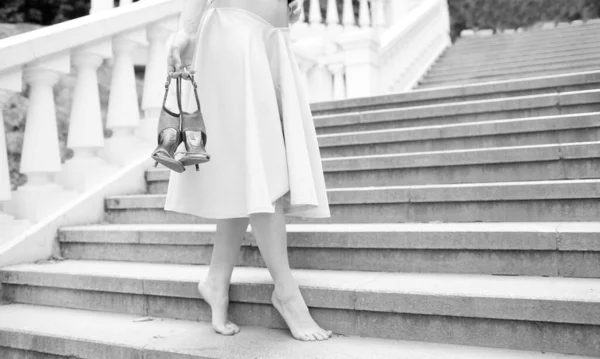 Young barefoot woman in dress stand on stepa alone. Tiptoe with pair of shoes on high heels in her hand. Posing alone. White and black picture. — Stock Photo, Image