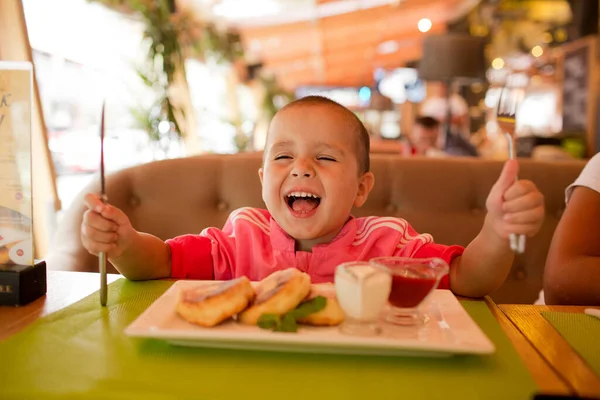 Young happy smiling girl have breakfast in opened cafe or restaurant. Sit at table and hold fork and knife by herself. Eating cheesecakes. — Stock Photo, Image