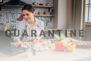 Neşeli yetişkin kadın mutfaktaki masada oturuyor. Klavyeye yazıp ekrana bakıyor. Kadın yemek yapıyor..