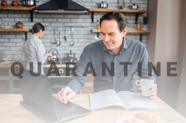 Neşeli yetişkin adam mutfaktaki masada oturuyor. Dizüstü bilgisayara bakar ve dokunur. Adam elinde bir fincan kahve tutuyor. Karısı sobanın başında duruyor..