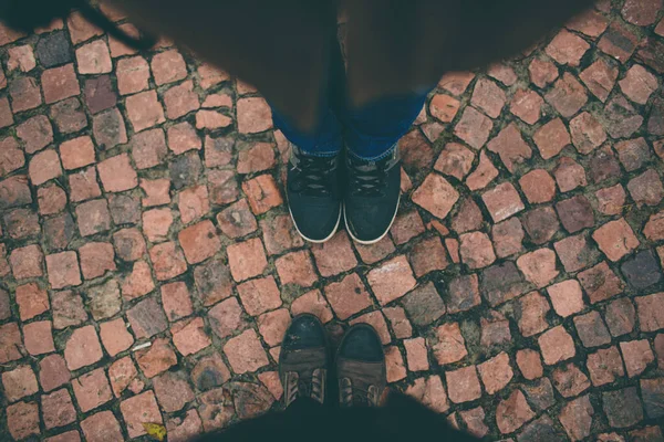 Blick von oben. Zwei Beinpaare stehen auf einer Straße aus Fliesen oder orangefarbenem Pflaster. Tragen Sie schwarze Hosen und Schuhe oder Turnschuhe. — Stockfoto