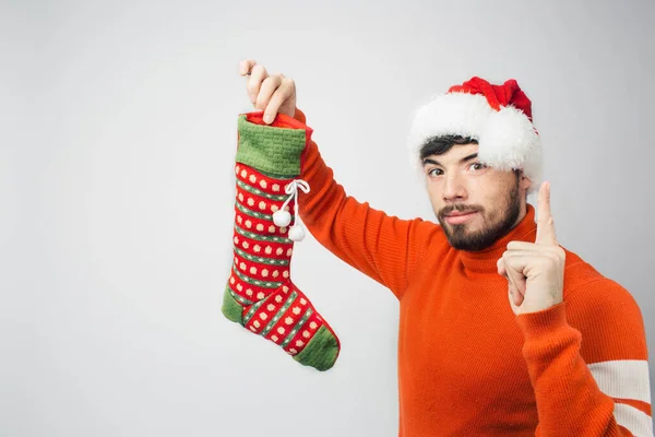 Jeune homme barbu isolé sur fond. Sérieux gars confiant montrer chaussette cadeau à la main. Pointez le doigt. Portez des vêtements rouges de Noël ou du Nouvel An. Père Noël aux cheveux foncés . — Photo
