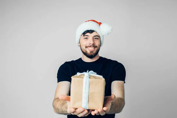 Jeune homme barbu isolé sur fond. Heureux gars gai positif dans un chapeau de Noël rouge festive tenir boîte cadeau ou cadeau dans les mains. Je pose devant la caméra. Seul en studio . — Photo