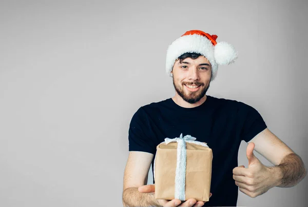 Jonge man met een baard geïsoleerd over de achtergrond. Man in zwart shirt en feestelijke rode kerstmuts poserend op camera. Houd grote duim omhoog en doos met cadeau of aanwezig in de hand. — Stockfoto