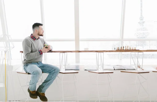 Young bearded man in cafe. Sits on high chair at long desk near window. Man holds glass with green drink and loos out window. On table chessboard. Laconic interior. — Stock Photo, Image