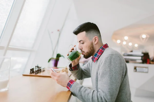 Young bearded man in cafe. He dressed in gray pullover and plaid shirt. Man is drinking tea and using smartphone. Chessboard, flower and blurred interior of cafe on background. — Stock Photo, Image