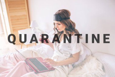 Young woman with dark hair sitting on bed in bedroom and working. Remote work in morning. Woman in gressing gown hold glass of red wine. Calm peaceful and concentrated.