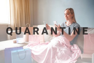 Evde online alışveriş. Tabletli ve akıllı telefonlu genç sarışın kadın, kanepede battaniyeyle otururken online dükkandaki malların fiyatlarını karşılaştırıyor.