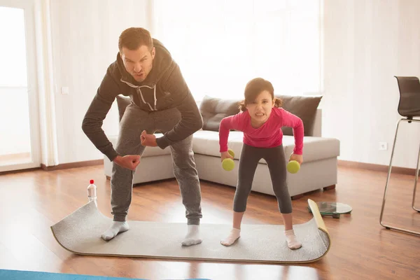 Père et fille s'entraînent à la maison. Entraînement dans l'appartement. Le sport à la maison. Ils font des visages et s'amusent sur un tapis de yoga avec des haltères . — Photo