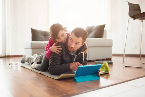 Far och dotter tränar hemma. Träning i lägenheten. Idrott hemma. Pappa är väldigt trött. Dottern klättrade på rygg och har kul. De använder en surfplatta och en sportapp — Stockfoto