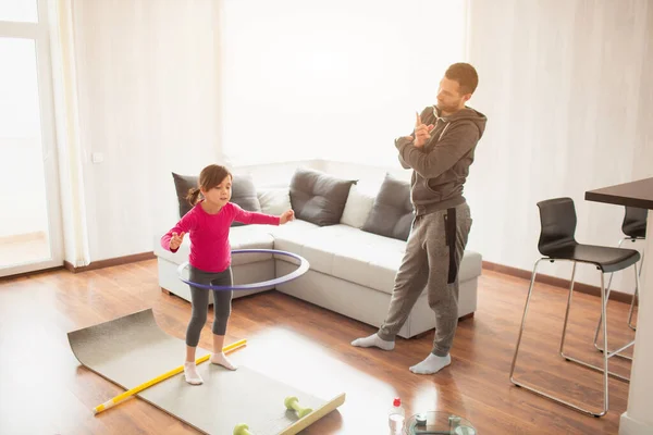 Батько і дочка тренуються вдома. Тренування в квартирі. Спорт вдома. вона накручує обруч і тато використовує спортивний годинник для вимірювання часу — стокове фото