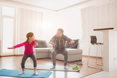 Baba ve kızı evde antrenman yapıyorlar. Apartman dairesinde egzersiz. Evde çömelme egzersizleri yapıyorum. Şirin çocuk ve babası odasının penceresinin yanındaki paspasın üstünde antrenman yapıyor.