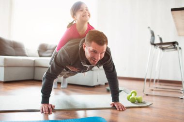 Baba ve kızı evde antrenman yapıyorlar. Apartman dairesinde egzersiz. Ev koşullarında spor. Tahtayı onlar yapıyor. Kızı babamın üstüne çıktı. Zeminden itiyor..