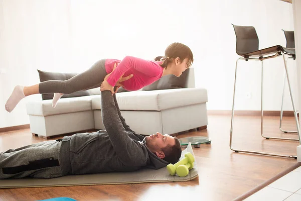Padre e hija están entrenando en casa. Entrenamiento en el apartamento. Deportes en casa. Papi yace en una esterilla de yoga, y sostiene a su hija en sus brazos — Foto de Stock