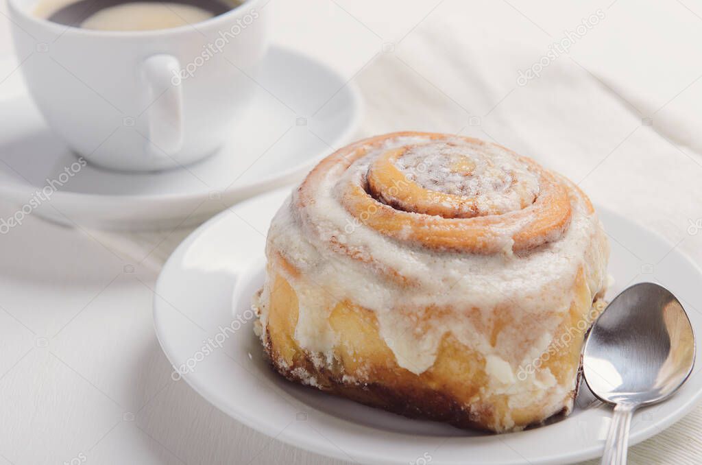 Hot cinnamon bun with sugar creamy icing on a white plate. Sweet breakfast or snack with a cup of coffee.