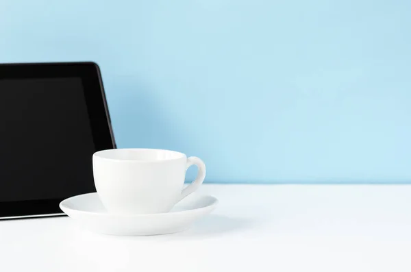 White work table with black tablet and white tea mug on a blue background. Copy space, minimal style.