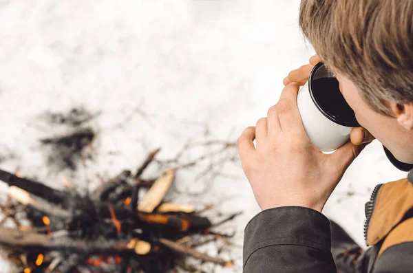 A man drinks coffee from a white mug by the burning campfire in the winter. Concept hike, walk, journey.