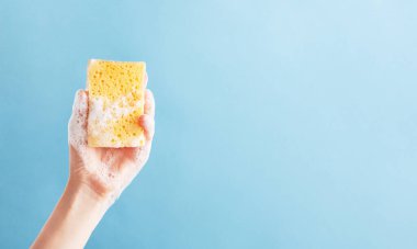 Bright yellow washcloth in woman hand. Sponge covered with shower foam isolated on blue background. Health beauty and hygiene concept. Copy space for text in right side. clipart